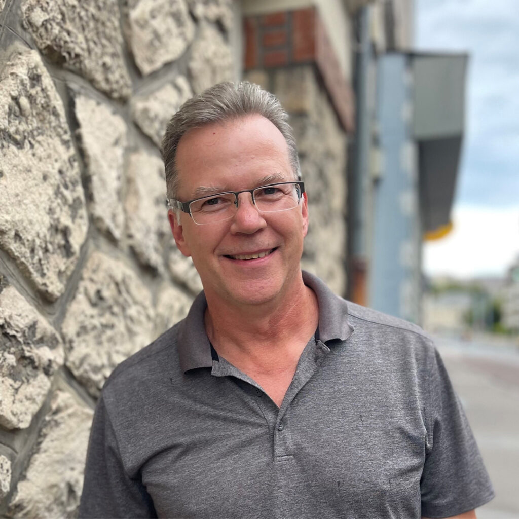 Stephen Parrott Man standing near stone wall of building with grey polo shirt and glasses - Stephen Parrott